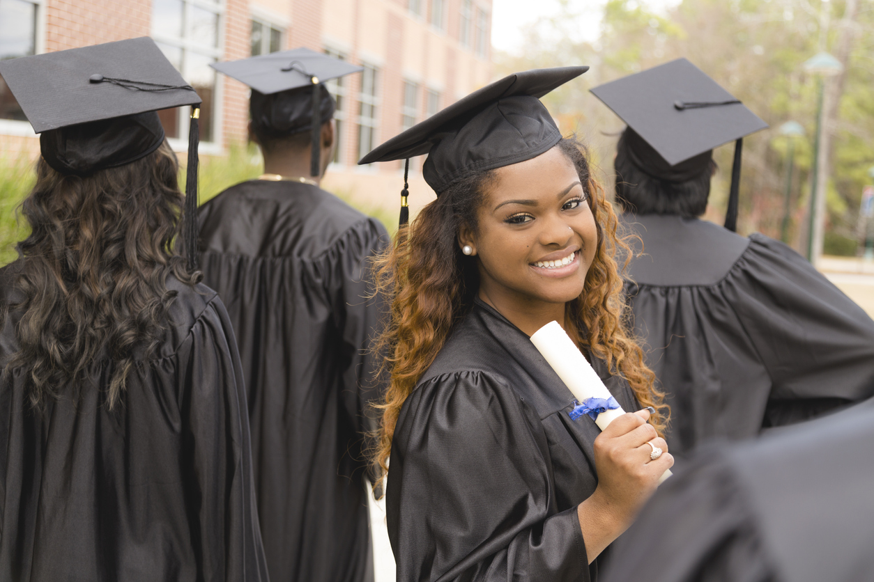 University or Liberal Arts College: Which is Right for You? Young woman in graduation robes holding college diploma on a college or university campus.