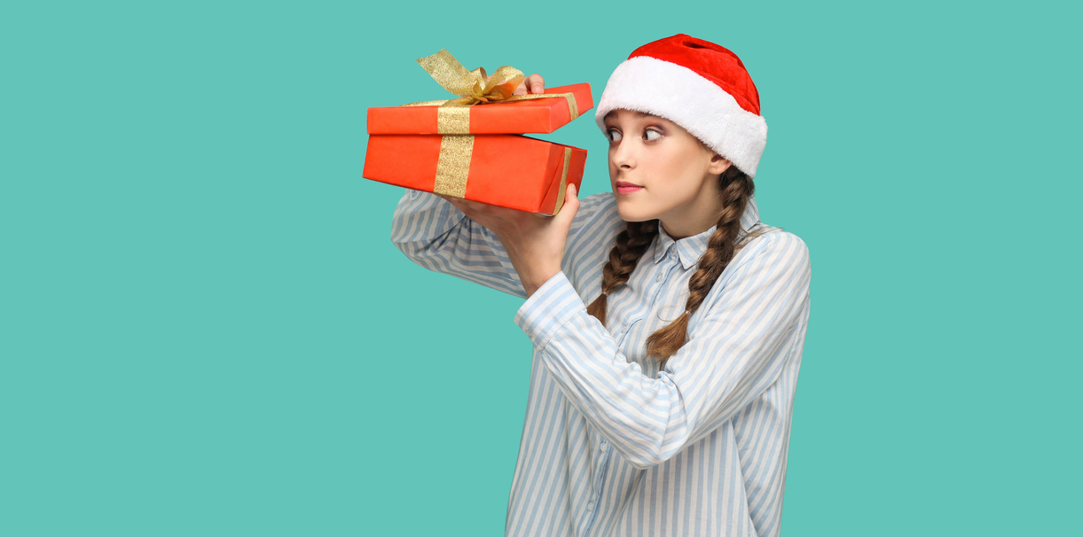 What Your High School Senior Really Wants for the Holidays beautiful girl in striped light blue shirt in red christmas cap standing holding red gift box, unboxing and looking inside